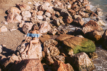 Luxurious blonde pose sitting on a rocky ocean in a beautiful blue swimsuit and a wreath of flowers on her head.