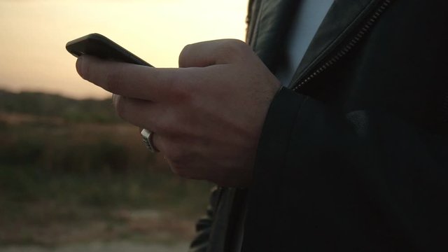 A man runs his finger on the screen of a smartphone against the backdrop of a sunny sunset, close-up. Background, slow motion