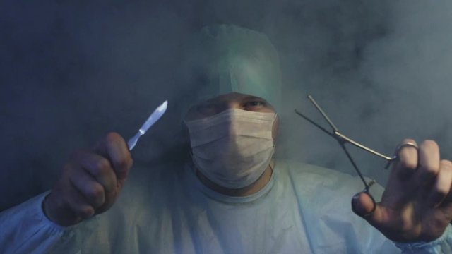 Surgeon with scalpel, grasper or forcep looking at the camera in smoke in slow motion. Doctor is ready to start cut a patient's tissues. Save a life or kill concept. Surgery and operating table