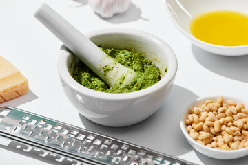 close up view of pesto sauce in pounder near grater, ingredients on white background