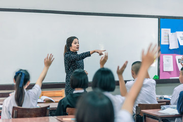A smiling Asian female high school teacher teaches the white uniform students in the classroom by asking questions and then the students raise their hands for answers.