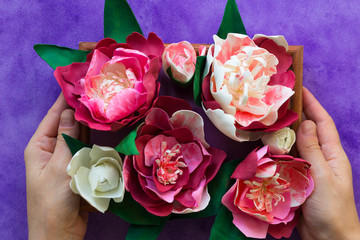 Handmade decorative peonies made from foamiran on wooden panel holding by woman hands on purple background