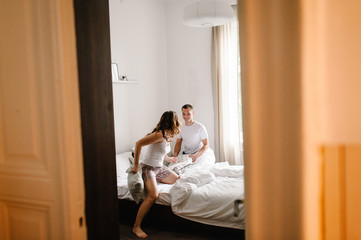 Obraz na płótnie Canvas Love couple beats pillows lying in bed, view through the door. A Satisfied and happy woman jumps with her husband. Emotions of happiness beautiful young couple. Lifestyle.