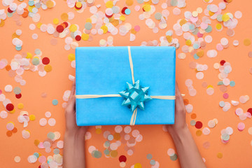 cropped view of woman holding blue present with bow near confetti on orange