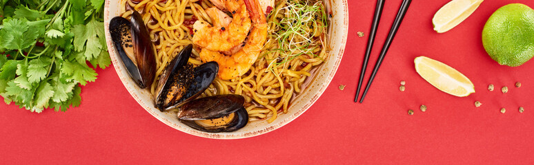 top view of spicy seafood ramen near chopsticks, parsley and lime isolated on red, panoramic shot
