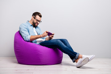 Full length profile photo of positive guy sitting comfy violet armchair holding telephone playing...