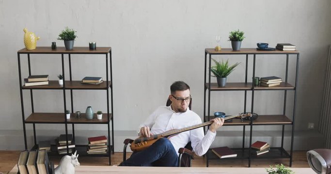 Man at work playing guitar for pleasure. Businessman is playing on guitar and enjoying it sitting in office during break. Guy practicing musical instrument. amera moves away from man. Music hobby.