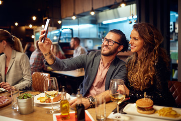 Cute caucasian smiling couple in love sitting in restaurant and taking selfie. Around them are their friends chatting and having dinner.