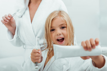 selective focus of cheerful kid holding toothpaste and toothbrush near mother