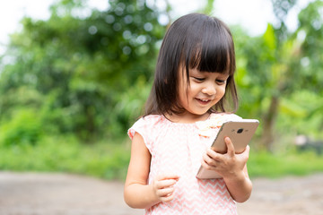 Adorable 3 years old asian little girl is laughing while she use the smartphone outside the house, concept of kid and technology using. 