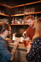 Happy caucasian couple leaning on bar counter and chatting with bartender. Pub interior. Nightlife. Focus on bartender.
