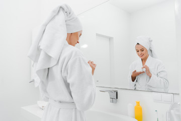 selective focus of happy woman holding container with cosmetic cream in bathroom