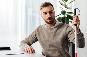 Visually impaired man in wireless earphones holding walking stick at table