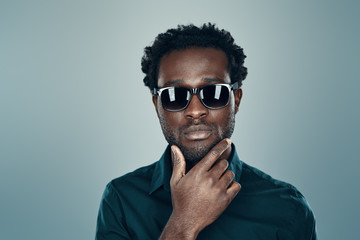 Obraz premium Handsome young African man in eyewear looking at camera and keeping hand on chin while standing against grey background