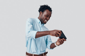 Handsome young African man playing mobile game and smiling while standing against grey background © G-Stock Faces