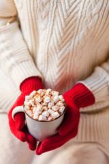 Young girl in knitted white wool sweater and red gloves is holding a mug with hot chocolate or coffee with marshmallow. Christmas lights on, cozy holiday atmosphere, aromatherapy for winter season. 