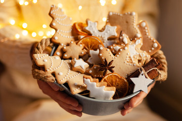 Young girl in knitted white sweater is holding a basket with homemade gingerbread cookies decorated with snow white icing. Anise and cinnamon as decor. Close up. Christmas lights on background