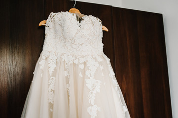 The bridal style vintage elegant dress with lace on a hangar on the background of a brown wooden cabinet. An expensive gipur. Bride's Preparations. Wedding Morning.