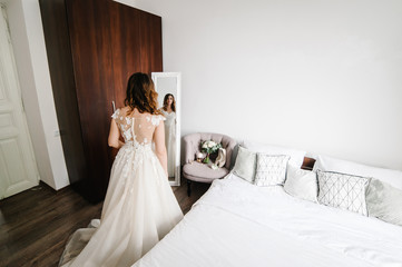 Fototapeta na wymiar Young attractive smiling bride in dress stand in front of the mirror and looking. Reflection. Portrait woman. The concept of the holiday and love. Wedding Morning Preparations. Rear view.