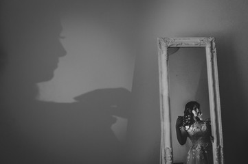 Artwork, noise, grain. Shadow of a woman. Portrait young attractive bride in dress stand in front of the mirror. Reflection body contour in the mirror on room. Rear view. Black and white photo.
