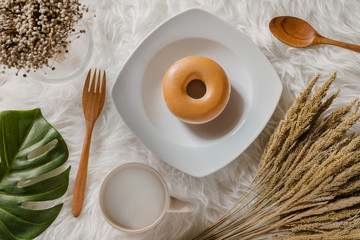 Tasty round doughnut on a white plate bordered with dry wheat grass, monstera green leaf and grass flowers in a glass jug with a mug of milk and wooden spoon and fork beside the plate on white fur rug © KsDear