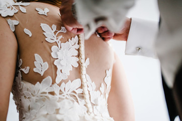 The man fastens buttons on the corset on the dress. Bride in white wedding dress with lace standing in the room.