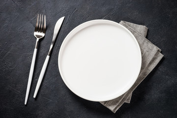 White plate, cutlery and napkin on black table top view.