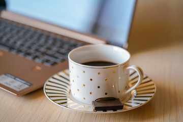 Cup of coffee on the wooden table with notebook on the background. Piece of black chocolate is on the saucer, break time in the office for refreshment