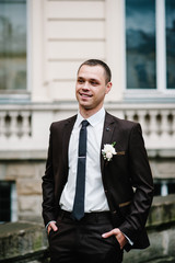 Attractive happy groom with boutonniere on wedding suit stand near old building outside, vintage palace outdoor.