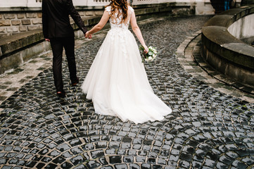 Fototapeta na wymiar The bottom of the bride's dress and groom's suit in old city. Newlyweds with a wedding bouquet of flowers go back to the palace on an old stone pavement.