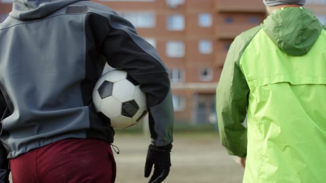 Back view following shot of two male athletes in sportswear walking together on soccer field with ball while going to train outdoors on autumn day