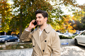 Young attractive man in trench coat happily talking on cellphone outdoor