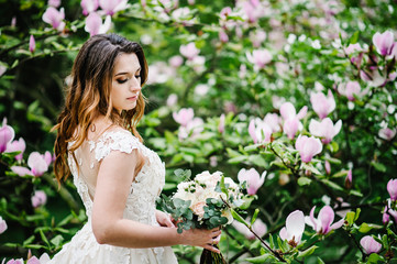 Fototapeta premium Portrait of bride with a wedding bouquet standing back on background nature of purple flowers of magnolia and greens. outdoors. looking down. upper half length.