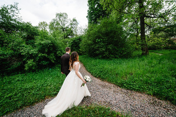 Romantic couple newlyweds, bride and groom is walking back on a trail in an green park. Happy and joyful wedding moment.