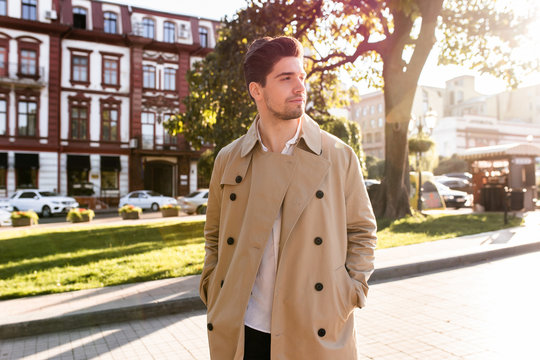 Young confident stylish man in trench coat intently looking away walking through street