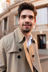 Young attractive stylish man in trench coat happily looking away outdoor