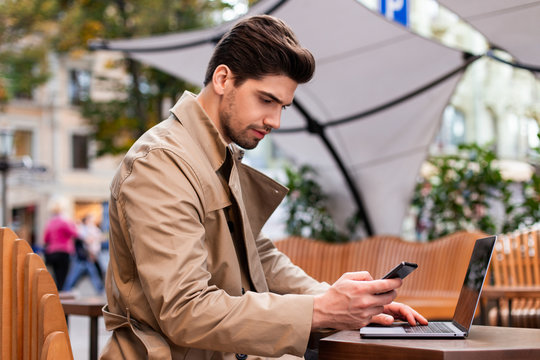 Young attractive businessman in trench coat with cellphone working on laptop in cafe on street