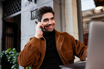Young cheerful man happily talking on cellphone working on laptop in cafe on street
