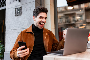 Young cheerful man working on laptop with cellphone and rejoicing in cafe on street