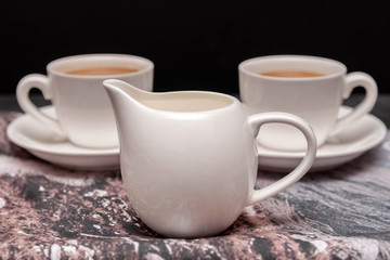 Mountain milk advertisement. White  milk creamer and two white coffee cups on gray napkin on black background