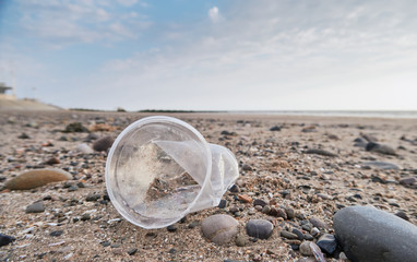 Fototapeta na wymiar Plastic litter cup and trash washed up on a sand and pebble beach. Environmental plastic pollution issue causing world news. Health disaster on shores across the world.