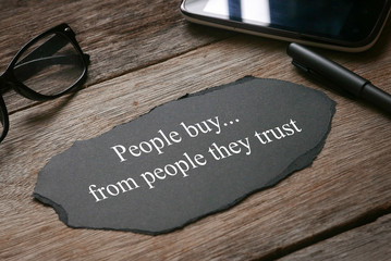Glasses,mobile phone,pen,a a piece of black paper written with People buy...from people they trust on wooden background.