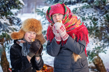 A boy in a scarf, furs coat and mittens together with an older girl in a red shawl  and warm mittens in the russian style eating pies on the backround of snow and forest.