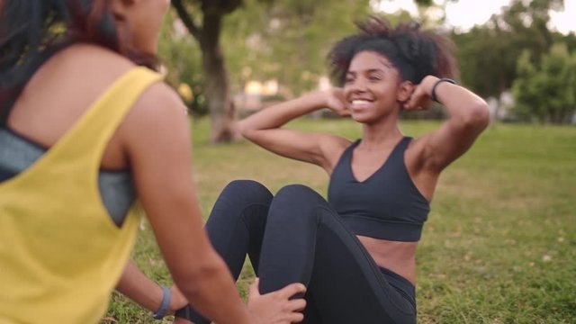 Young woman doing sit-ups with assistance of her female friend in the park - friends doing exercise in the park supporting eachother