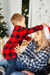 Little boy puts on a Christmas Santa hat to mom. Celebrating Christmas and New Year, having fun, happy time together.