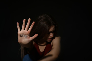 Abused young woman showing palm with word STOP against black background, focus on hand. Domestic...