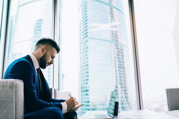 Lumped businessman in stylish suit writing notes in day planner with laptop at office