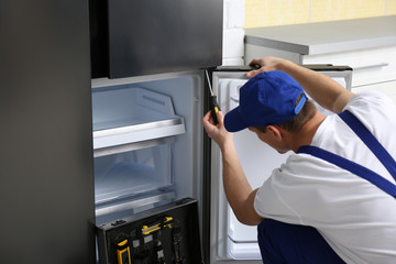 Male technician with screwdriver repairing refrigerator in kitchen