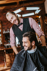 Fototapeta na wymiar handsome barber in latex gloves putting hand on shoulder of happy man in barbershop