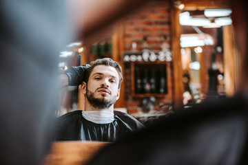 selective focus of barber styling hair of bearded man near mirror in barbershop